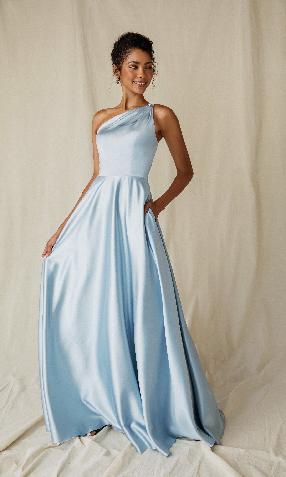 A woman stands before a cream fabric backdrop wearing the Eureka 9025, a light blue one-shoulder long satin formal dress.
